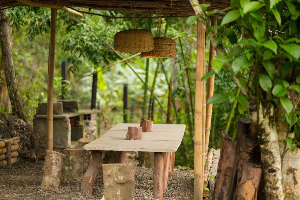 Zona de comedor rústica al aire libre en Rivabella Eco Lodge, con mesa de cemento, bancas de troncos y techo de bambú en medio del bosque.